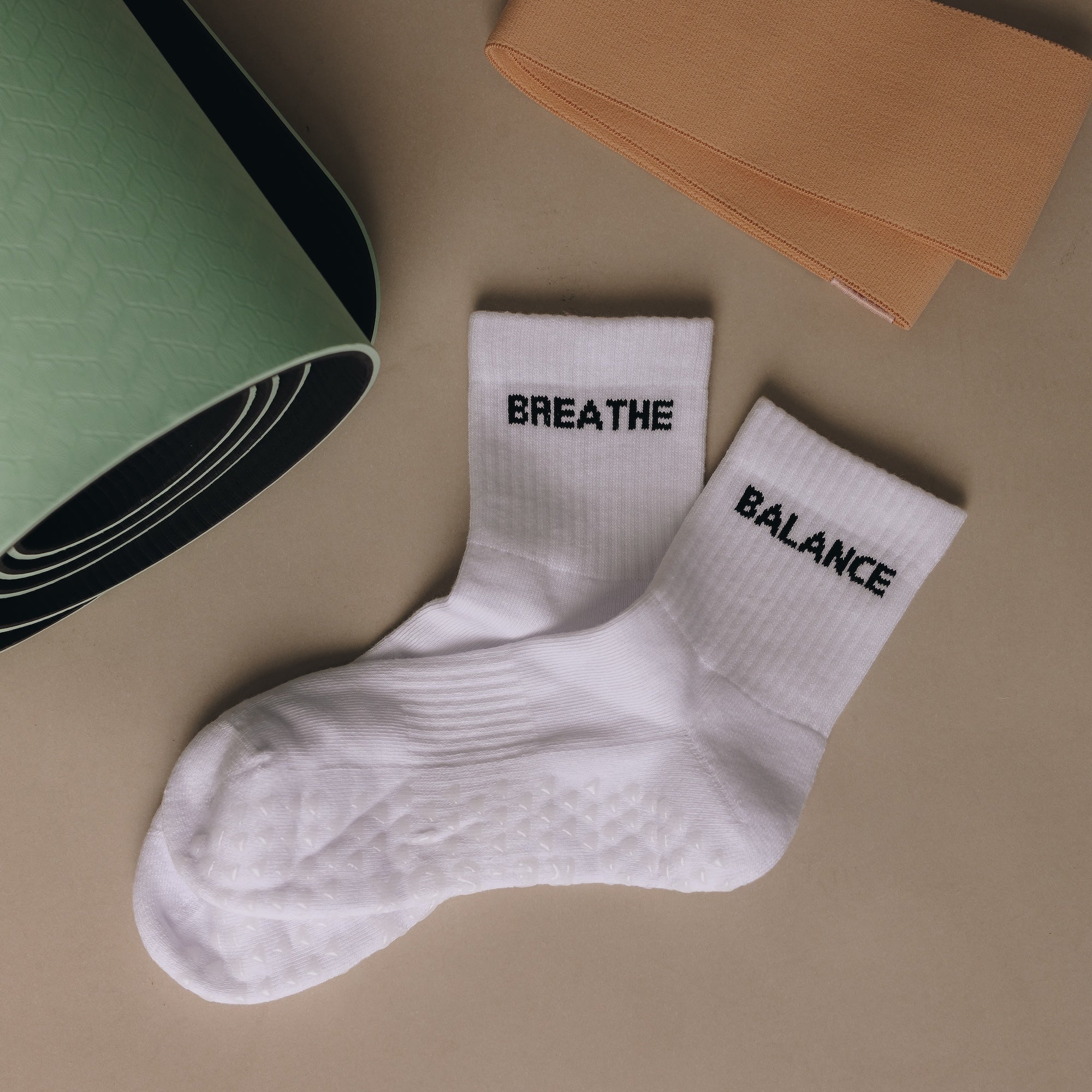 White grip socks with 'BREATHE' and 'BALANCE' text on a beige surface with a yoga mat and book.