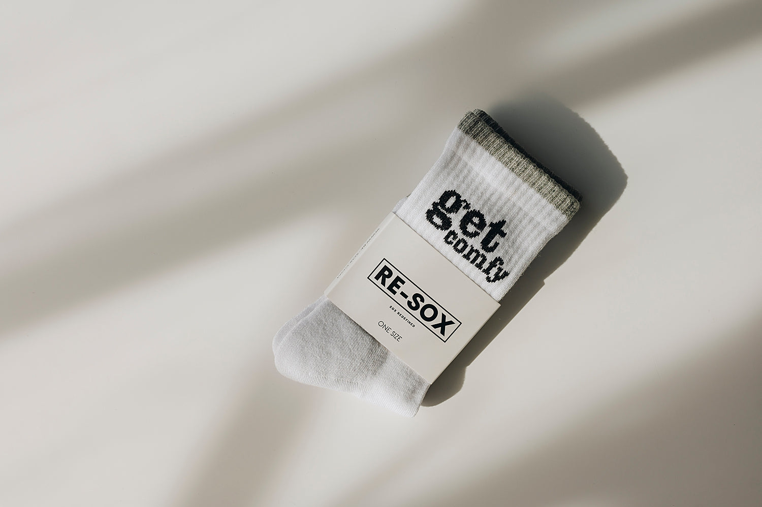 A pair of white ribbed crew socks with the text 'get comfy' printed on the side and light grey stripe around the top, packed in white sock card with RE-SOX logo on the front. 